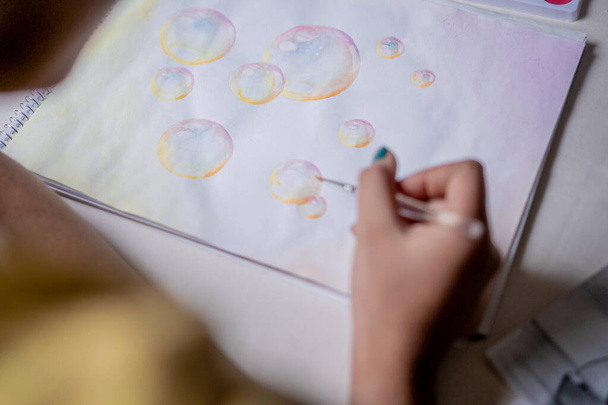Жінка малює барвистий малюнок бульбашки в ескізі з кольоровими акварелями на білому столі
. - Фото, зображення