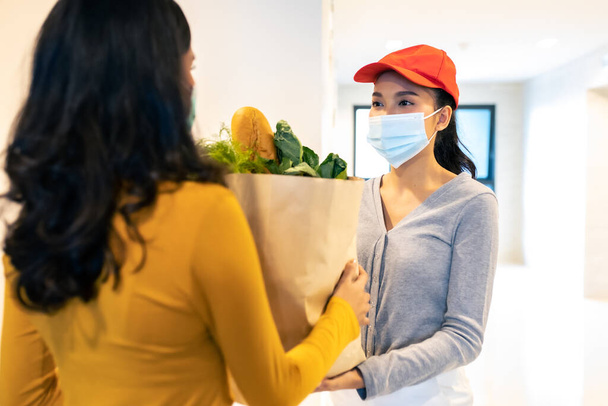 Asian woman face mask at home and deliver woman wearing face mask handling bag of food, fruit, vegetable give to female costumer in front of apartment.  grocery delivery service during covid19. - Foto, imagen