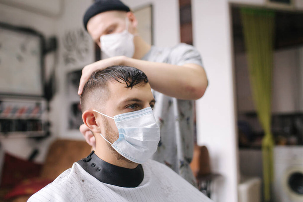 Un uomo che si fa tagliare i capelli da una forbice in barbiere. Il barbiere usa forbici e maschera durante la pandemia di coronavirus. Il barbiere professionista lavora a casa. Quarantena Covid-19 - Foto, immagini