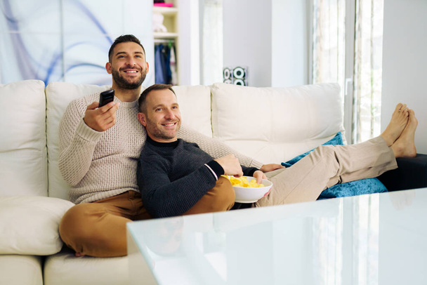 Gay couple sitting on the couch at home watching something on TV and having a snack. - Foto, afbeelding