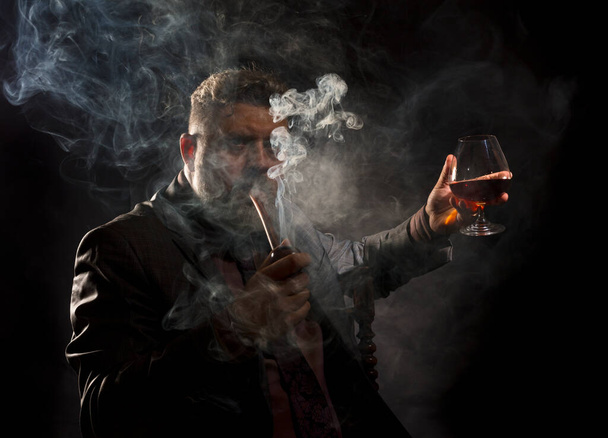 Solid confident bearded man in suit with glass of whisky smoking pipe - Photo, image