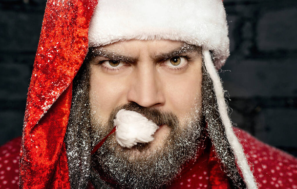 Close-up portrait of a young santa claus in a red sweater and a red cap with a black beard against a black wall - Foto, immagini