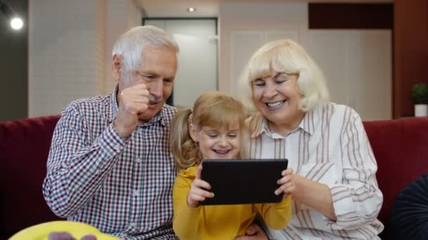 Senior Caucasian couple with cute child girl granddaughter using tablet, watching funny videos - Footage, Video