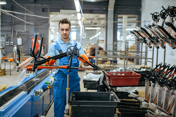 Bicycle factory, worker holds black teen bike frame. Male mechanic in uniform installs cycle parts, assembly line in workshop, industrial manufacturing - Photo, Image