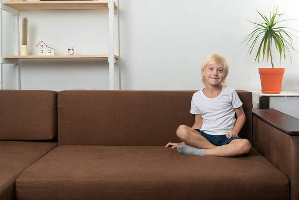 Szőke tinédzser fiú, aki keresztbe tett lábbal ül otthon a kanapén. Boldog fiú. Fénymásolási hely. - Fotó, kép