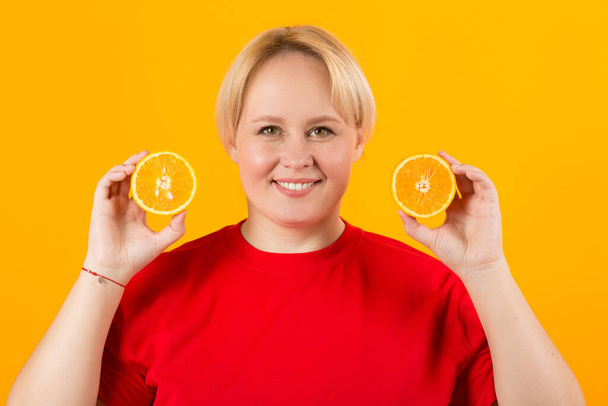 beautiful young plump female in a red t-shirt on a yellow background with oranges - Φωτογραφία, εικόνα