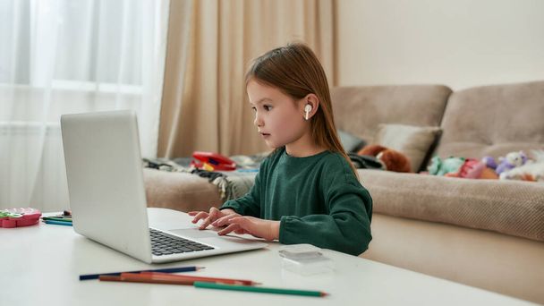 A little girl enjoying her time with a laptop being in a huge bright guestroom during distance education alone - Foto, afbeelding