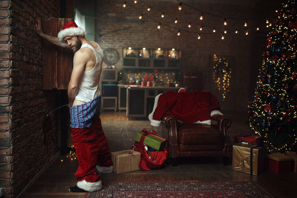 Schlecht angetrunkener Weihnachtsmann mit rotem Hut, der an die Wand uriniert, fiese Party, Humor. Ungesunder Lebensstil, bärtiger Mann im Urlaubskostüm, Silvester und Alkoholismus - Foto, Bild