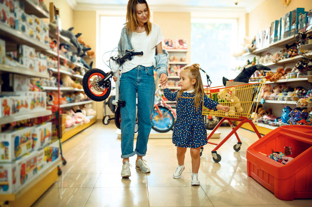 Mother and pretty little girl buying bicycle in kids store. Mom and adorable daughter near the showcase in toyshop, happy childhood, family makes a purchase in shop - 写真・画像