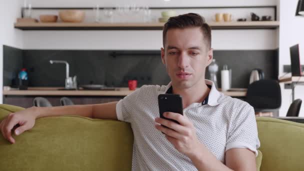 Закрыть жизнерадостного молодого человека, сидящего на диване с помощью смартфона, улыбкой в современной квартире текстового сообщения прокрутки технологии прослушивания - Кадры, видео