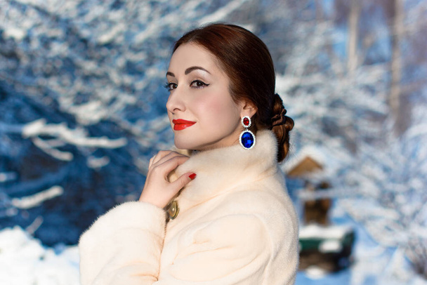 Winter beauty, portrait of fashionable gorgeous woman. Fairy Christmas in the snow-covered forest. The girl in a fur coat with accessories ruby gold earrings. Beautiful elegant lady with red lips and bright make up in a frosty day - Photo, Image