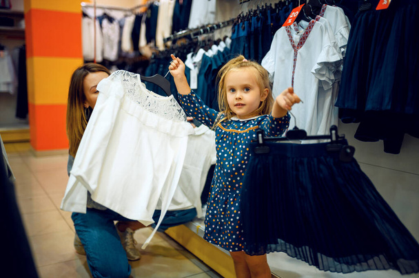 Mother and little baby choosing skirt in kids store. Mom and adorable girl near the showcase in childrens shop, happy childhood, family makes a purchase in kids market - Zdjęcie, obraz