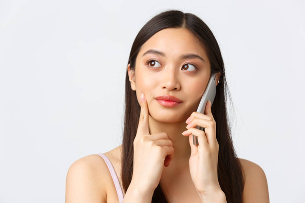 Beauty, fashion and people emotions concept. Close-up portrait of pretty asian woman having phone call, looking away thoughtful, booking table in cafe, deciding what order, talking on smartphone - Photo, image