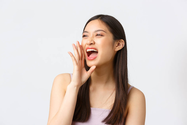 Beauty, fashion and people emotions concept. Close-up portrait of carefree happy asian girl making loud announcement, holding hand near opened mouth as shouting, white background - Foto, immagini