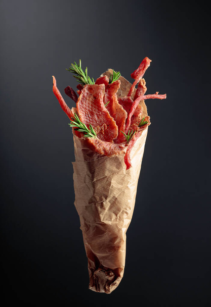 Snijdt gekruid gedroogd vlees met rozemarijn in een papieren zak op een donkere achtergrond. - Foto, afbeelding