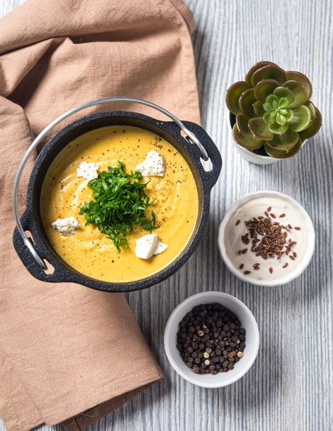 A pumpkin puree soup in a cast iron bowl with white cheese chunks and chopped parsley on top of a grey wooden table. Beige napkin cloth, white cream sauce with flax seeds, a bowl of black peppercorns. - Photo, Image