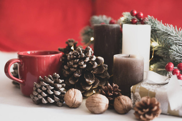  Різдвяний декор. Червоний кухоль з кавою, свічки, кедрові шишки, гілка хвойного дерева, різдвяний подарунок на червоному тлі
. - Фото, зображення