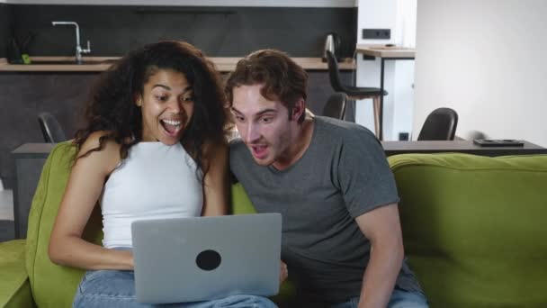 Szczęśliwa wielorasowa młoda para korzystająca z laptopa do zakupów online razem oglądając film wideo siedzący na kanapie w salonie. Uśmiechnięty biały mężczyzna i afrykańska amerykańska kobieta patrzą na komputer w domu. - Materiał filmowy, wideo