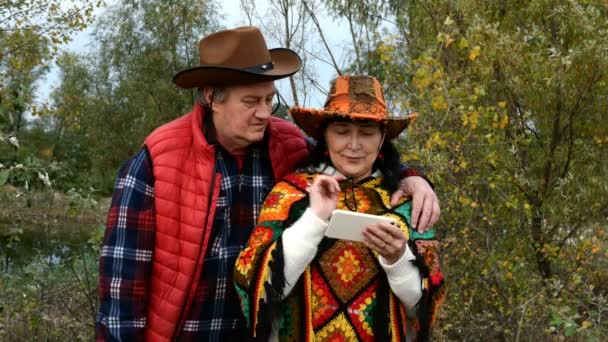 Felnőtt, középkorú, cowboy kalapos pár, akik futárral beszélgetnek, vagy nézegetnek valamit az okostelefonjukon. Az őszi parkban. Közelkép. - Felvétel, videó