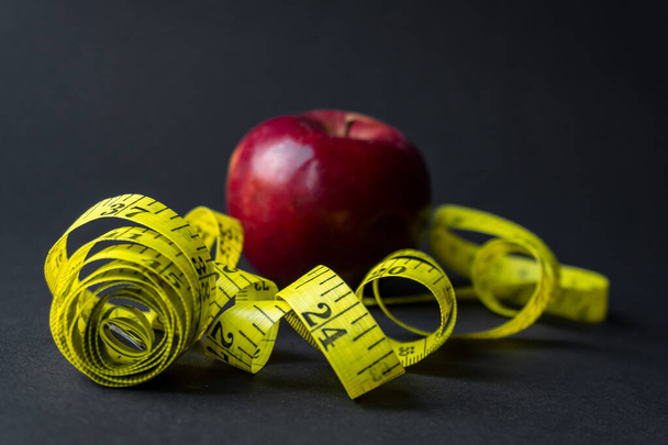 Ein roter Apfel, in ein flexibles gelbes Zentimeterband gewickelt, liegt darauf. dunkler Hintergrund. gesunde Ernährung, Vitamine und Mineralstoffe. - Foto, Bild