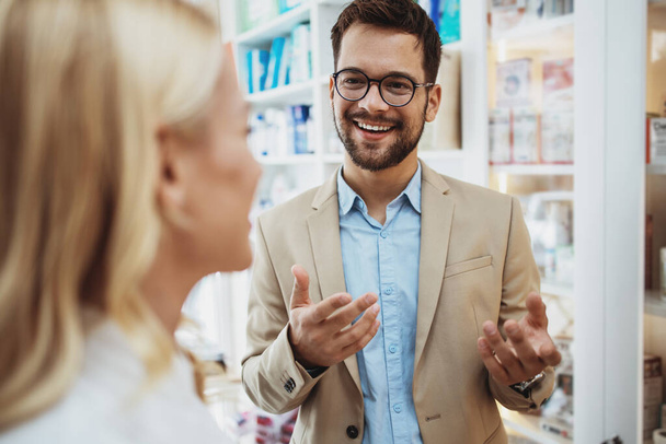 Young business man choosing and buying drugs in a drugstore while talking with attractive female pharmacist. She helping him with expert advices. - Foto, imagen