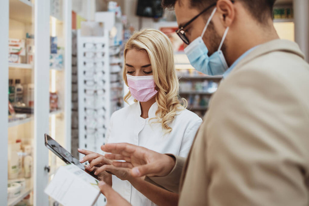 Young business man choosing and buying drugs in a drugstore while talking with attractive female pharmacist. She helping him with expert advices. They are wearing protective face masks against virus infection. - 写真・画像