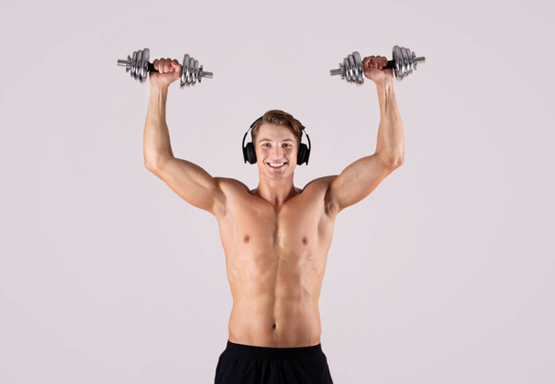 Sportlicher junger Mann mit nacktem Oberkörper, der Hanteln hebt und Musik über Kopfhörer auf hellem Studiohintergrund hört - Foto, Bild