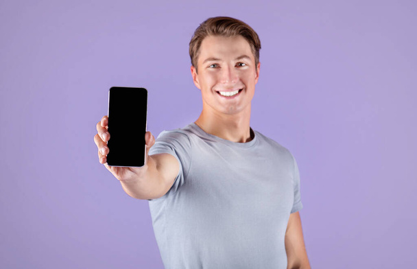 Feliz joven deportista mostrando teléfono inteligente con pantalla vacía sobre fondo lila, maqueta para el diseño de aplicaciones móviles - Foto, imagen