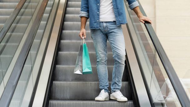 Nahaufnahme von Männerbeinen in Jeans mit Einkaufstüten, während er am schwarzen Freitag auf der Rolltreppe im Einkaufszentrum steht - Foto, Bild