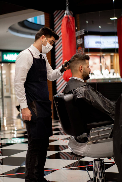 ευρύ πλάνο ενός κομψού κουρέα με μάσκα κόβει τα μαλλιά ενός πελάτη με καυκάσια χαρακτηριστικά - Φωτογραφία, εικόνα