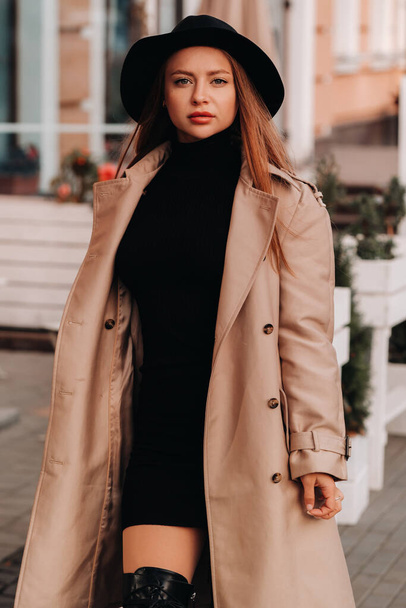 Stylish young woman in a beige coat in a black hat on a city street. Women's street fashion. Autumn clothing.Urban style. - Fotografie, Obrázek