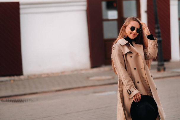 Stylish young woman in a beige coat and a black hat in his hands and glasses on a city street. Women's street fashion. Autumn clothing.Urban style - Foto, Imagen