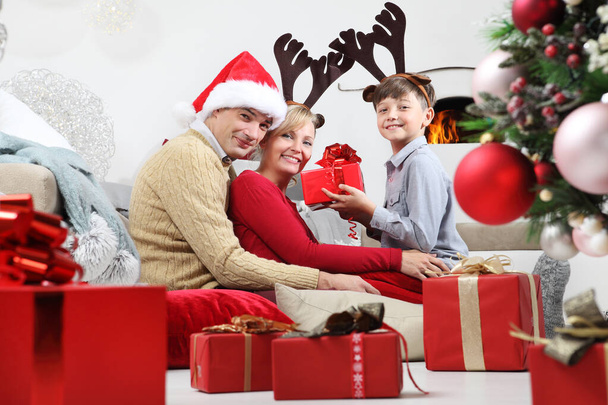 feliz Navidad, feliz estancia familiar en casa, padres dando el regalo a su hijo cerca del árbol iluminado y decorado, alegre y sonriente sentado en la sala de estar y consolado por la chimenea - Foto, imagen