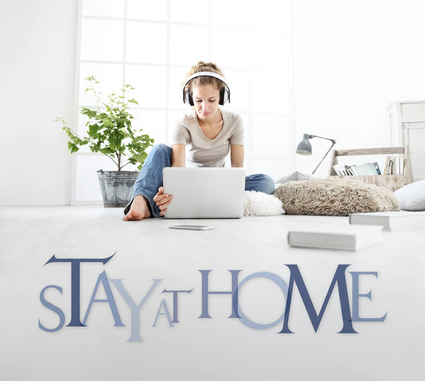 young woman with computer and headphones, look laptop monitor, with stay at home text isolated on white floor background, stay at home social media campaign for coronavirus covid-19 prevention - Photo, Image