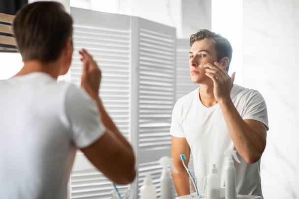 Handsome guy looking in the mirror applying cream on face - Photo, Image