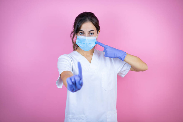 Νεαρή μελαχρινή γιατρός κορίτσι φορώντας νοσοκόμα ή χειρουργική στολή με γάντια λάτεξ και μάσκα πάνω από απομονωμένο ροζ φόντο που δείχνει τη μάσκα. Προειδοποιητική έκφραση με αρνητική και σοβαρή χειρονομία στο πρόσωπο με το χέρι στο στόμα γιατί βήχει. - Φωτογραφία, εικόνα