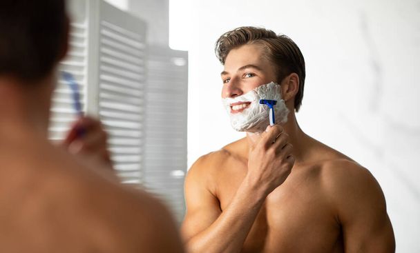 Handsome young guy looking in the mirror and shaving - Photo, Image