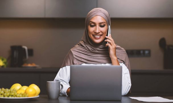 Mujer árabe feliz en el ordenador portátil hablando por teléfono en la cocina - Foto, imagen