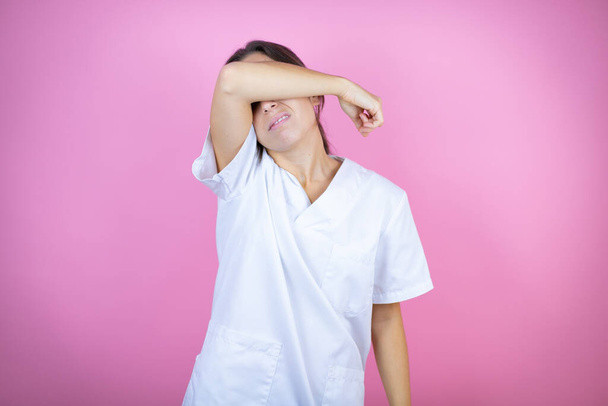 Молодая брюнетка доктор девушка, носящая медсестру или хирурга униформу на изолированном розовом фоне закрывая глаза с рукой улыбается веселый и смешной. Слепая концепция. - Фото, изображение