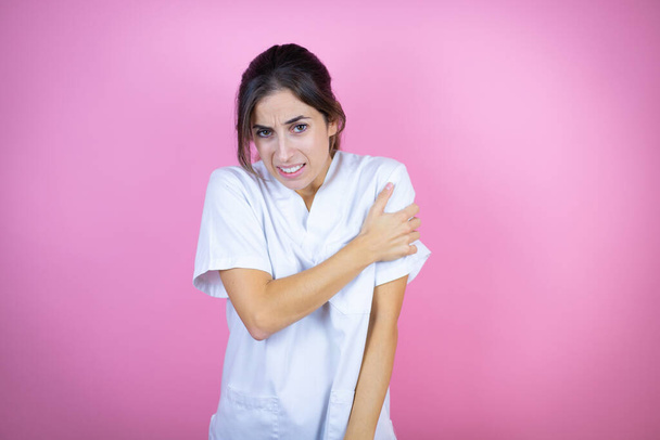 Junge brünette Ärztin trägt Krankenschwester- oder Chirurgenuniform über isoliertem rosa Hintergrund mit Schmerzen an der Schulter und einem schmerzhaften Ausdruck - Foto, Bild