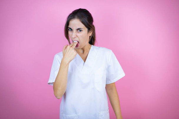 Young brunette doctor girl wearing nurse or surgeon uniform over isolated pink background disgusted with her hand inside her mouth - 写真・画像
