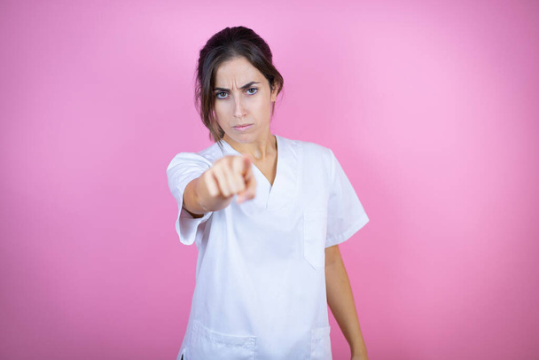 Mladá brunetka lékař dívka na sobě zdravotní sestra nebo chirurg uniforma přes izolované růžové pozadí ukazuje na přední s prstem - Fotografie, Obrázek