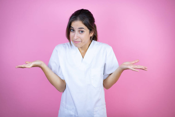 Nuori brunette lääkäri tyttö yllään sairaanhoitaja tai kirurgi yhtenäinen yli eristetty vaaleanpunainen tausta tietämätön ja sekava ilme kädet ja kädet nostetaan - Valokuva, kuva