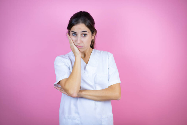 Молода брюнетка лікарка в уніформі медсестри або хірурга над ізольованим рожевим тлом думає, що виглядає втомленою і нудною з схрещеними руками
 - Фото, зображення