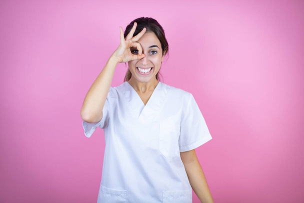 Young brunette doctor girl wearing nurse or surgeon uniform over isolated pink background doing ok gesture shocked with smiling face, eye looking through fingers - Valokuva, kuva