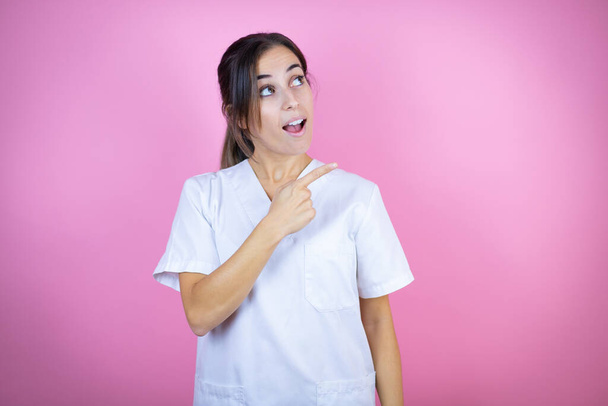 Joven doctora morena vistiendo uniforme de enfermera o cirujano sobre fondo rosa aislado sorprendida y señalando con la mano y el dedo hacia un lado - Foto, imagen