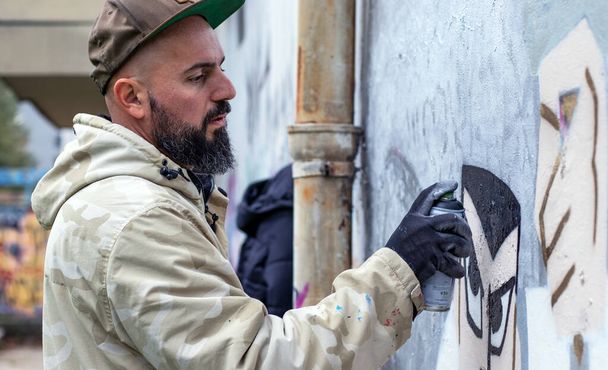 Street graffiti artist in action, drawing  with spray paint can on the wall in rainy weather. Street art culture concept.  - Photo, Image