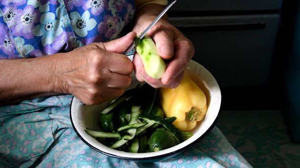 80 éves nő ráncos kezei, amint zöld héjat hámoz nyers uborkából konyhakéssel. Az idős hölgy kezei növényi ételt készítenek a rusztikus konyhában. - Fotó, kép