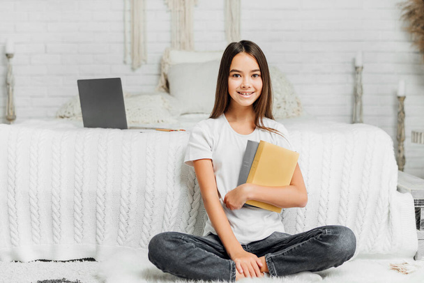 Happy child, beautiful girl with books, ready for online lesson, on the background of the bed in his room. - Foto, afbeelding