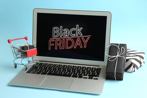 Laptop with Black Friday announcement, small shopping cart and gifts on light blue background - Фото, изображение